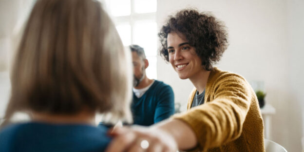 A woman smiling and reaching out to comfort another person in a supportive setting, reflecting compassion, connection, and emotional encouragement during recovery.
