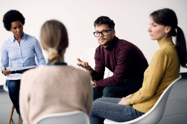 A group of people sit in a circle during a therapy session, with one man gesturing as he speaks.