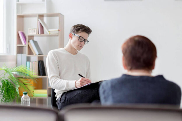 A young man in glasses writes on a clipboard while a person sits with their back to the camera.